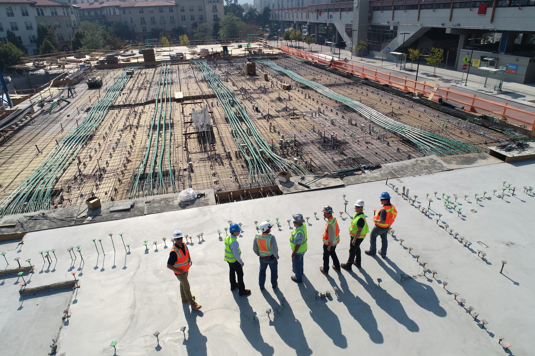 Seven workers in hard hats and high-visibility vests walking across a concrete deck above a rebar mat