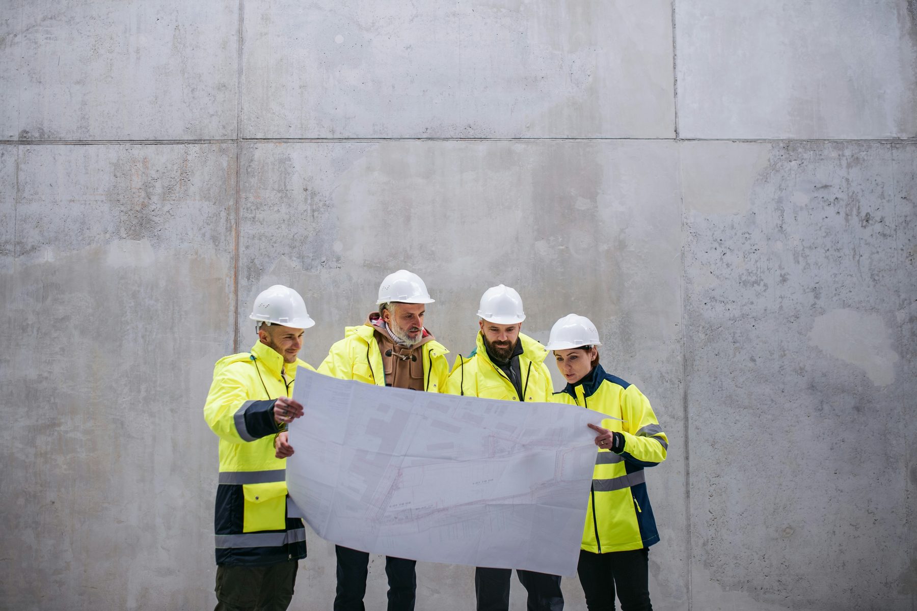 Four site personnel in high-visibility jackets and white hard hats reading a large drawing together against a concrete wall