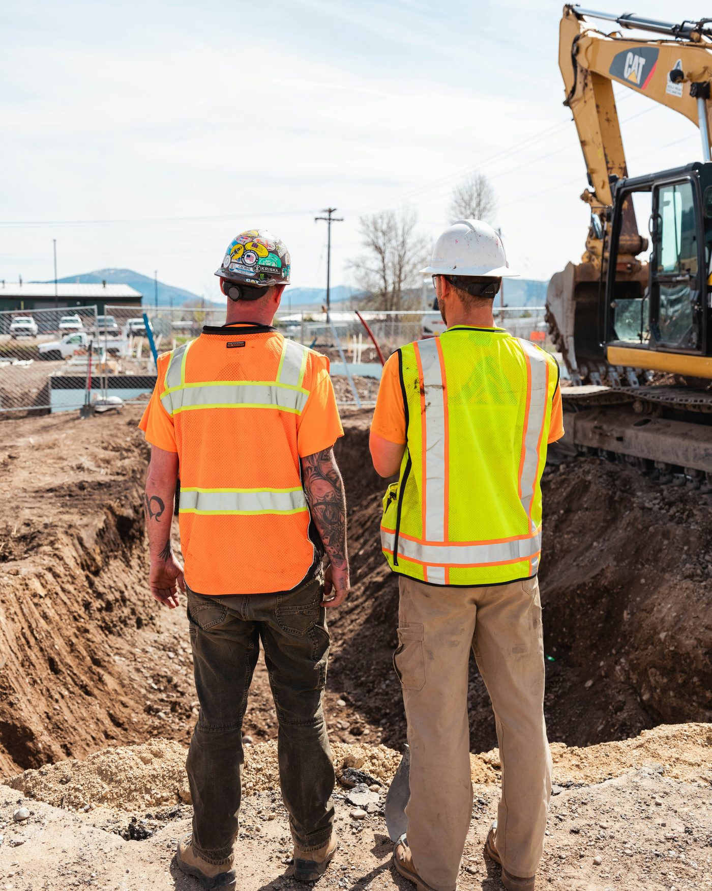 Two workers in high-visibility vests and hard hats standing at the edge of an excavation, watching an excavator work