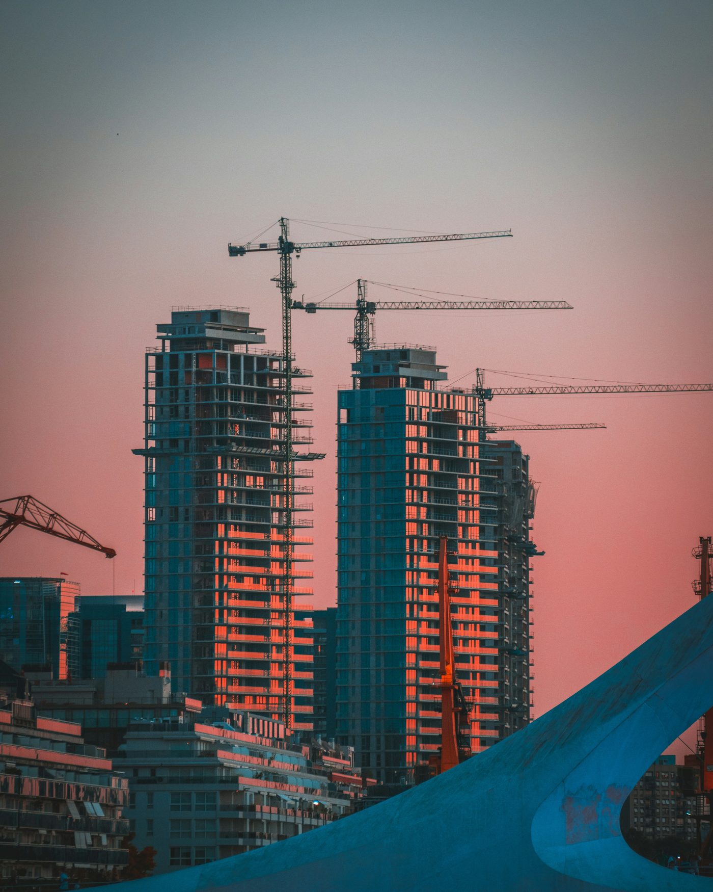 Two residential towers under construction with tower cranes above them at sunset