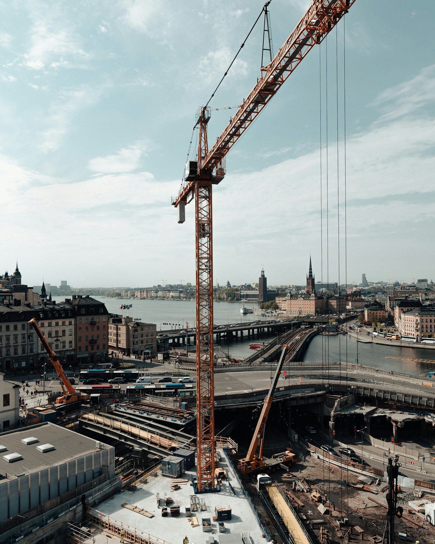 A tower crane over an excavated urban construction site beside a waterway, with workers on the deck below